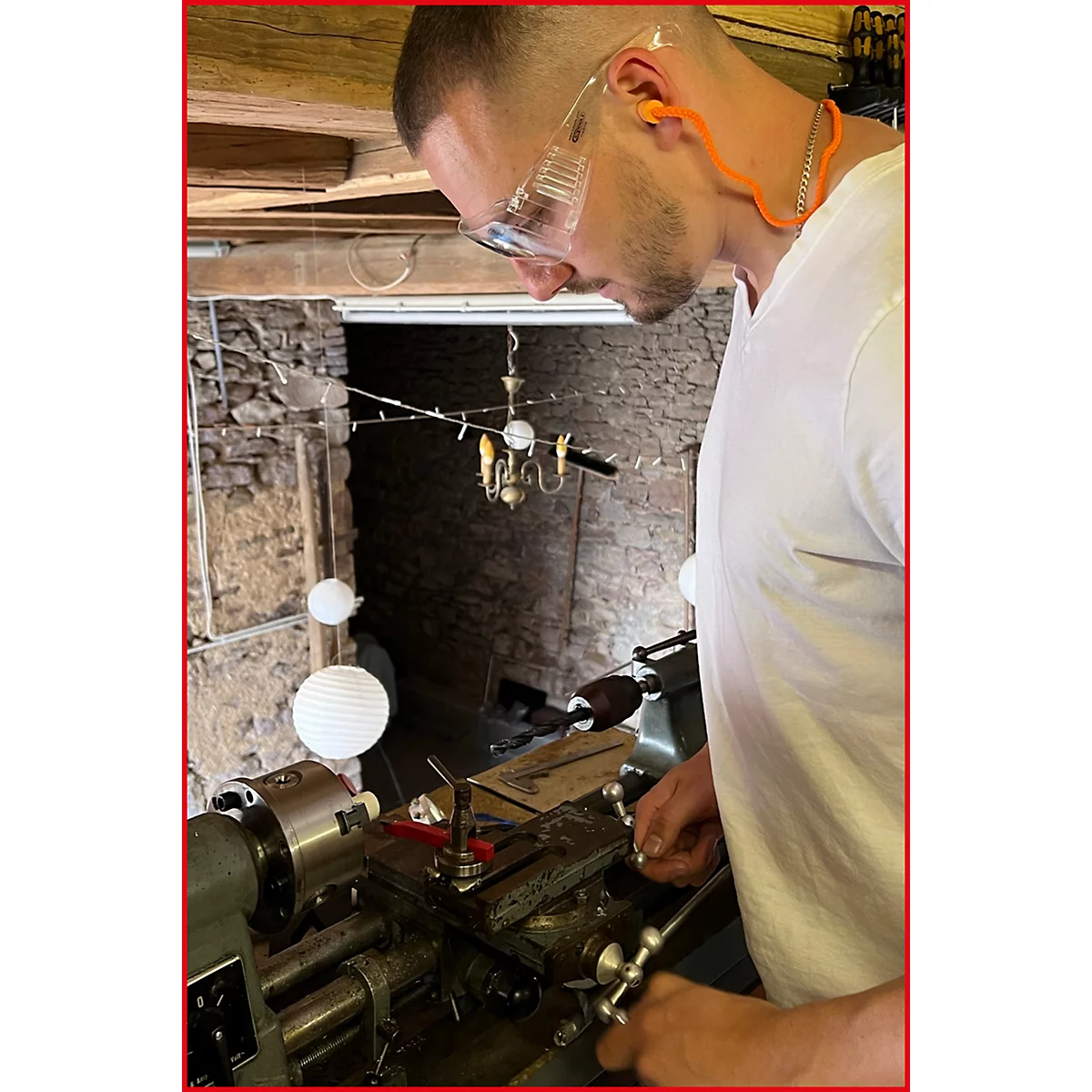 Mann mit Schutzbrille arbeitet an einer Drehmaschine, trägt Gehörschutz. Im Hintergrund: Steinmauer und Kronleuchter.